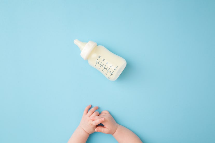 Infant hands and bottle of milk on light blue table background. Feeding time. Pastel color. Closeup....