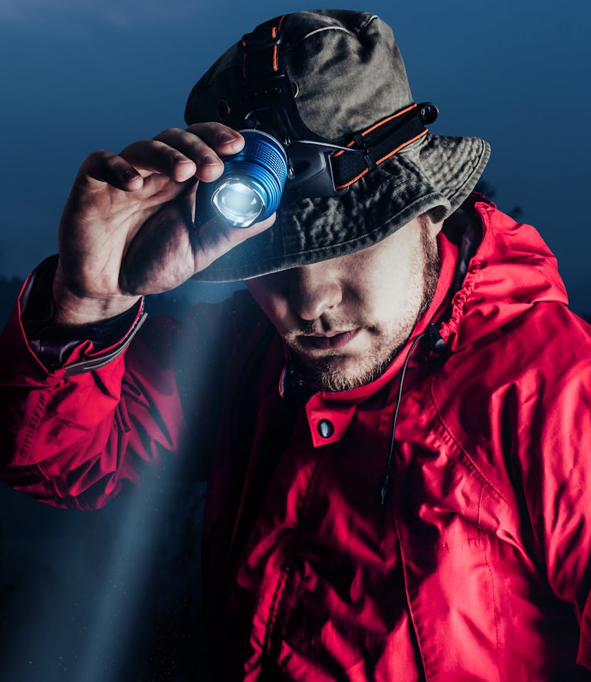 Photo of a male hiker in red jacket and panama putting on head mount flashlight on dark mountain for...