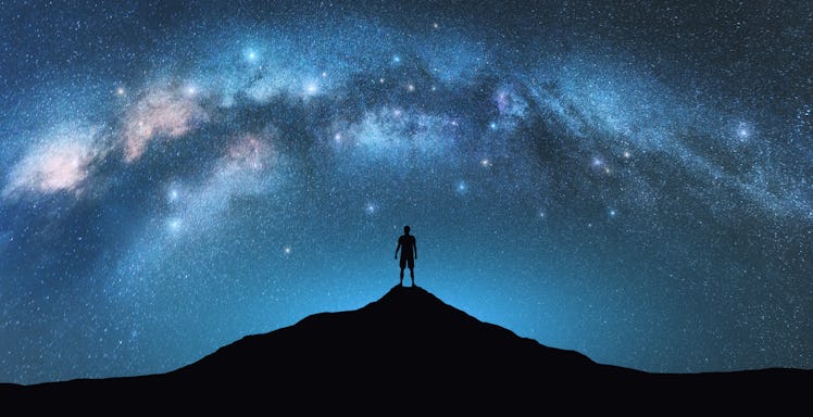 Milky Way arch and man on the mountain peak at starry night. Silhouette of alone guy, blue sky with ...