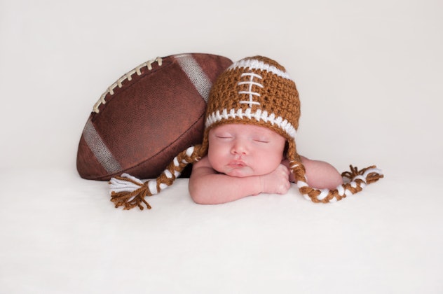 Portrait of a two week old, sleeping newborn baby boy. He is posed with an American football wearing...
