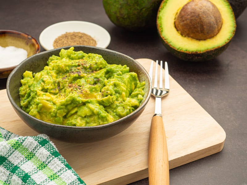 Fresh guacamole on a dish placed on a cutting wooden board with ingredients for homemade guacamole: ...