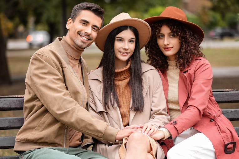 Man and two beautiful women sitting on bench in park. Polyamory concept