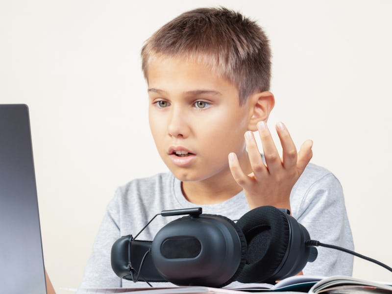 Shocked annoyed upset child with laptop computer and headphones sitting at table with books. Educati...