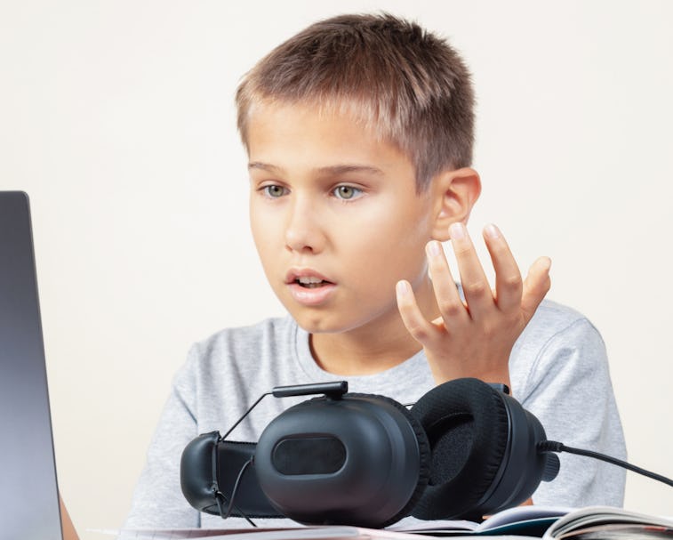 Shocked annoyed upset child with laptop computer and headphones sitting at table with books. Educati...