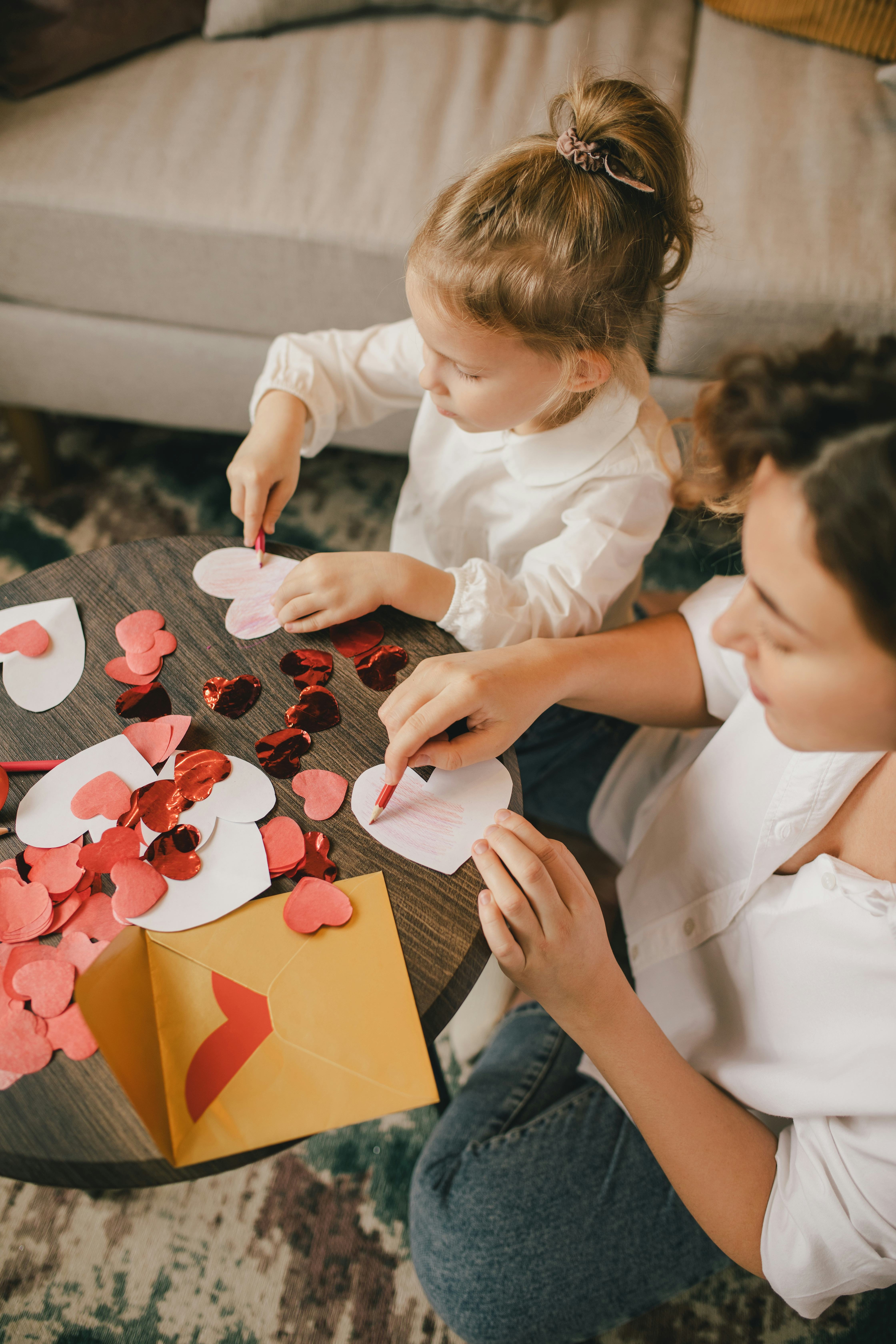 Mother and little daughter making Valentine's day cards using color paper, scissors and pencil, sitt...