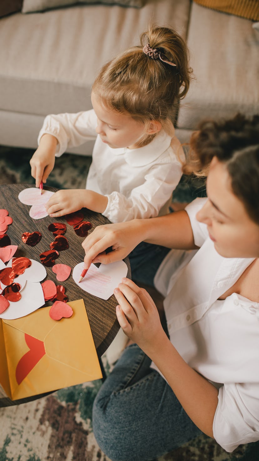 Mother and little daughter making Valentine's day cards using color paper, scissors and pencil, sitt...