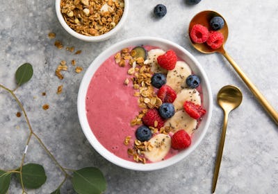 Summer acai smoothie bowls with raspberries, banana, blueberries, and granola on gray concrete backg...