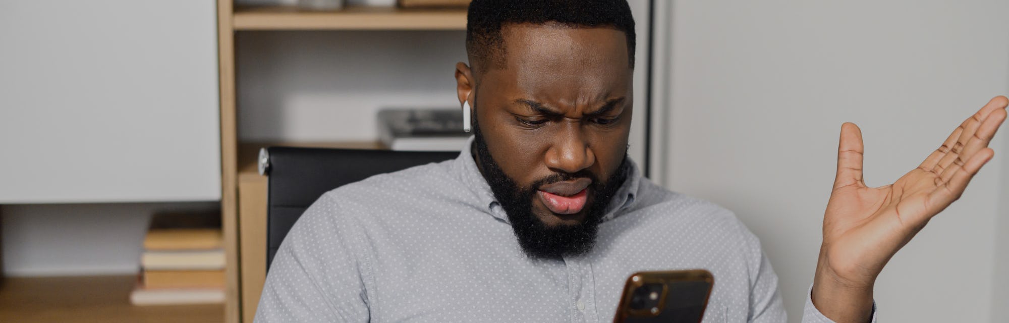 Confused puzzled African-American man in smart casual shirt holding smartphone and expressing misund...