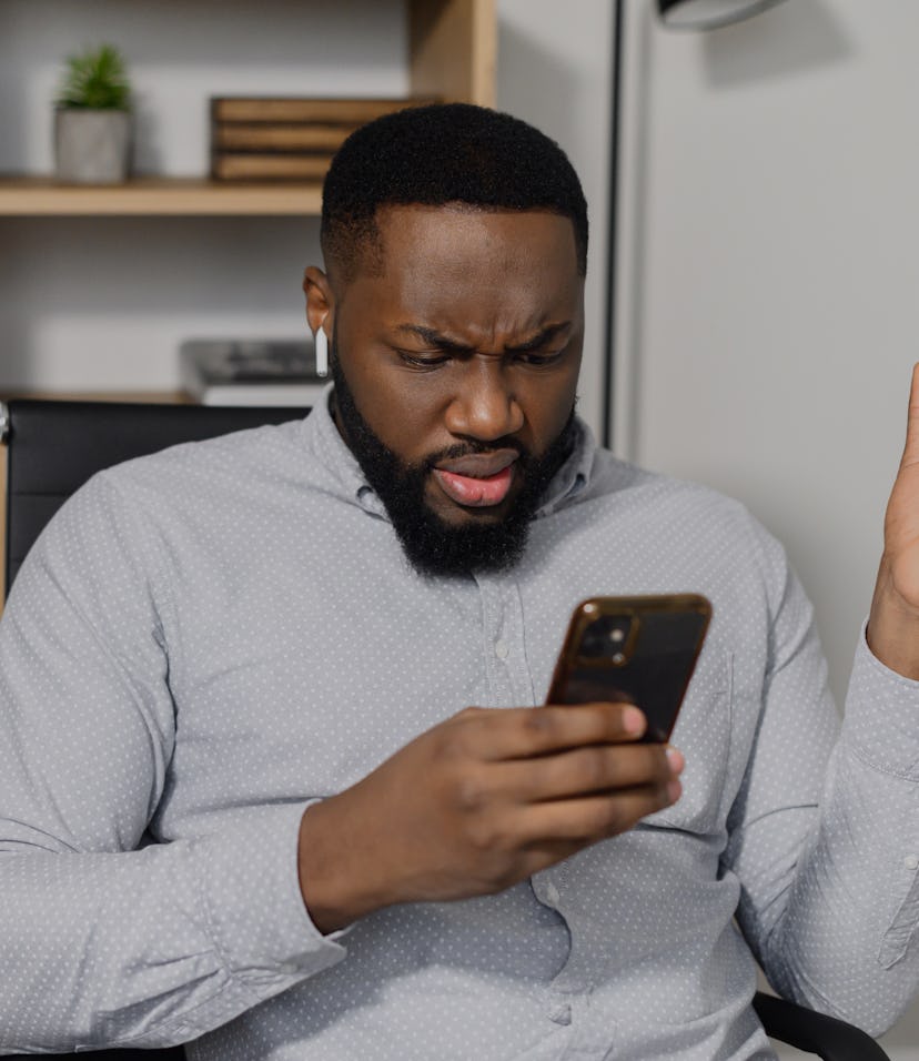 Confused puzzled African-American man in smart casual shirt holding smartphone and expressing misund...