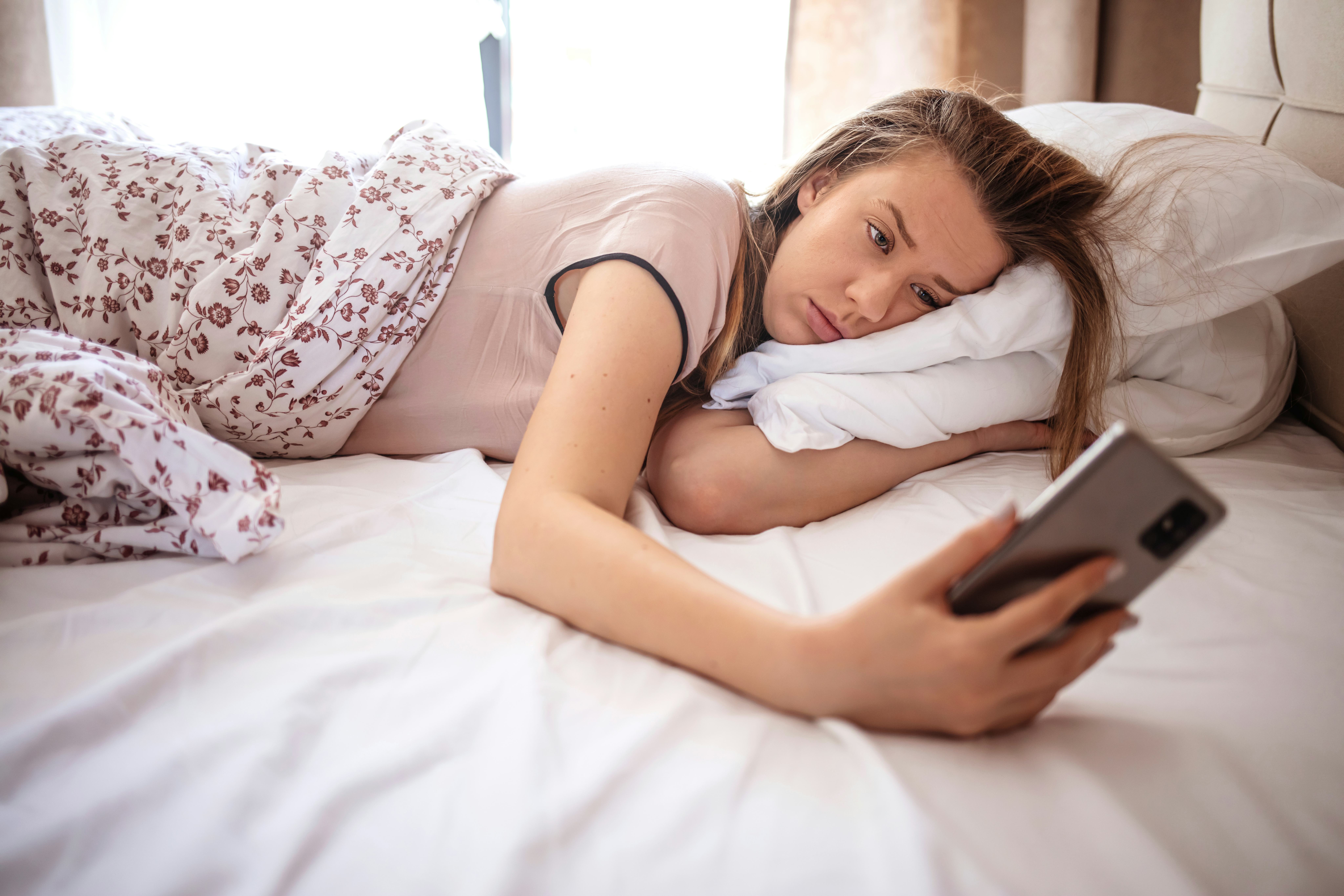 Woman lying in bed reaching to turn off the alarm on her mobile phone in morning. Sleepy woman being...
