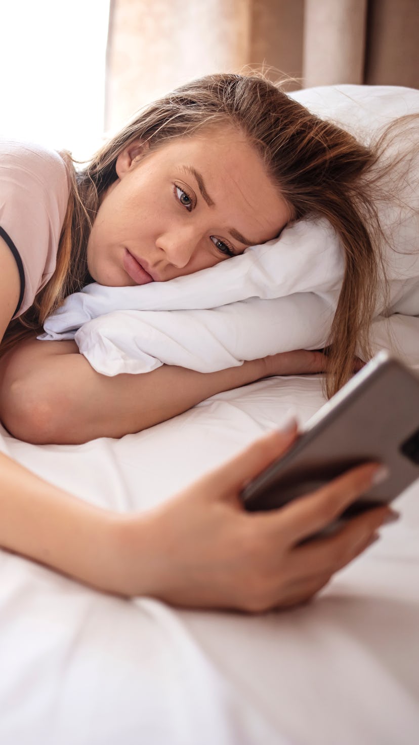 Woman lying in bed reaching to turn off the alarm on her mobile phone in morning. Sleepy woman being...
