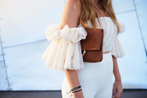 Street style image of young woman wearing an off the shoulder frill white top, white trousers and ch...