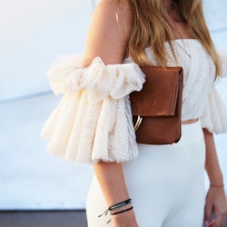 Street style image of young woman wearing an off the shoulder frill white top, white trousers and ch...