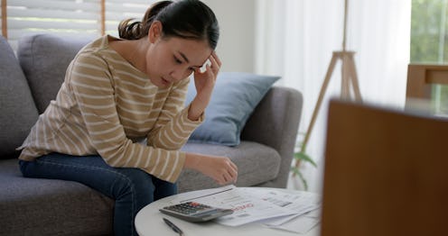 Upset tired young asia woman sit on sofa at home think hard worry in tax expense saving issue past d...