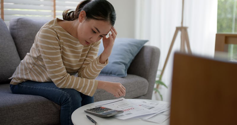 Upset tired young asia woman sit on sofa at home think hard worry in tax expense saving issue past d...