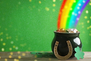 Pot with gold coins, horseshoe and clover leaves on wooden table against green background, space for...