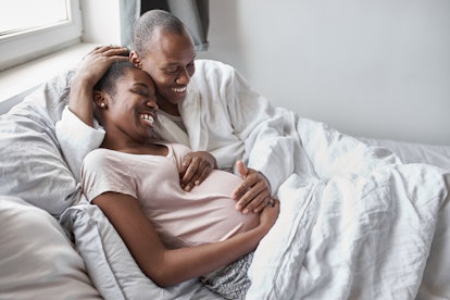 Young Black husband cuddling pregnant Black woman on bed, both smiling widely. Heart rate increases ...