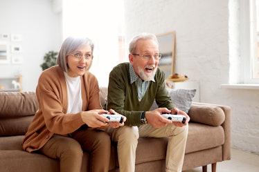 Excited concentrated senior couple husband and wife playing video console games together while sitti...