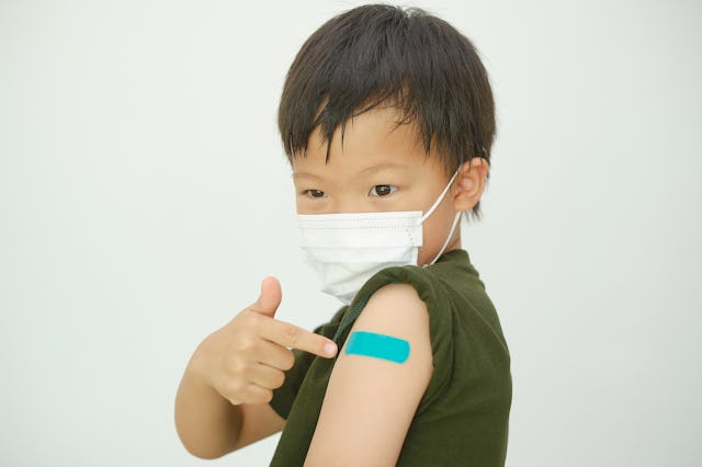 Doubtful Young child wearing medical mask showing his arm with bandage after receiving Covid vaccine