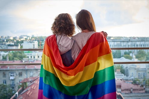 Two girls with rainbow flag looking out the window at the city. Lesbian couple celebrating pride mon...