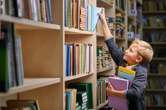 school boy taking books from shelves in library, with a stack of books in hands. child brain develop...