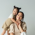 cheerful asian toddler daughter hugging happy mother isolated on gray