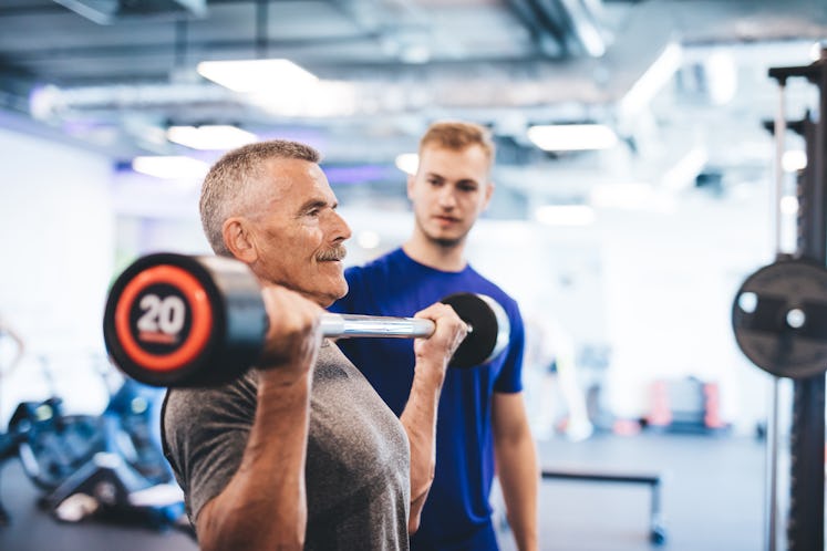 Older man lifting weights, supervised by gym assistant. Sporty lifestyle of elderly people.