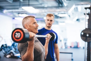 Older man lifting weights, supervised by gym assistant. Sporty lifestyle of elderly people.