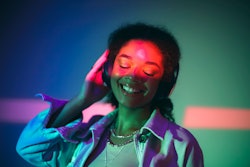 A woman is listening to headphones and looking joyful under neon-colored lights. The Jupiter-Neptune...