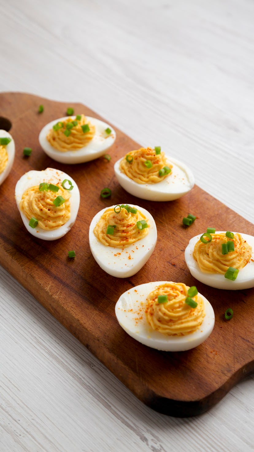 Homemade Deviled Eggs with Chives on a rustic wooden board on a white wooden background, side view. ...