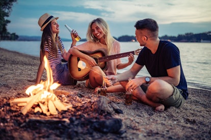 Group of cheerful friends playing the guitar and having fun on the beach by the bonfire, for which t...