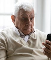 Old man sit on sofa hold smartphone look at device screen feels confused shocked by received sms mes...
