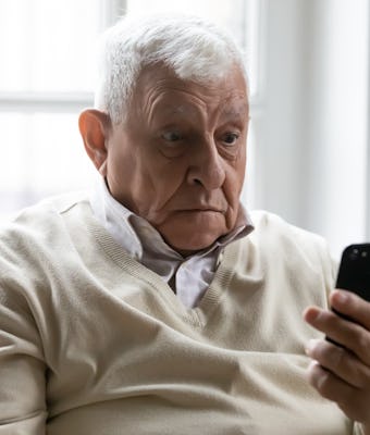 Old man sit on sofa hold smartphone look at device screen feels confused shocked by received sms mes...