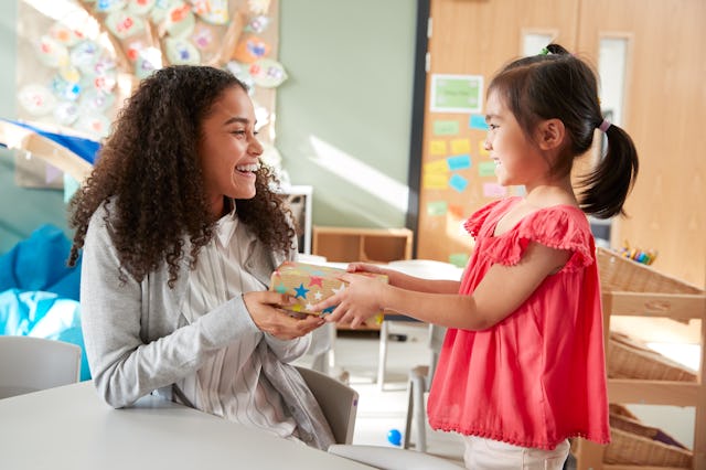 Kindergarten schoolgirl giving a gift to her female teacher in a classroom, side view, close up