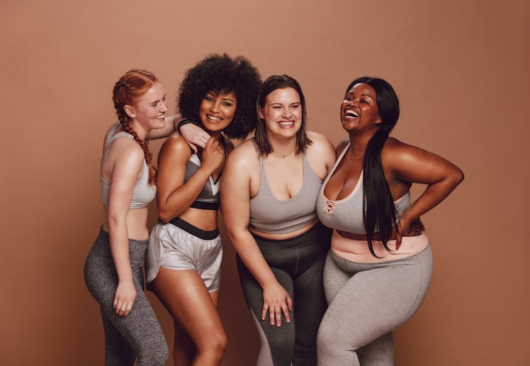 Smiling group of women in different size standing together in sportswear against brown background. D...