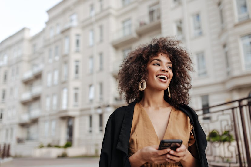 Cheerful African woman holds smartphone. Curly brunette lady in brown blouse and black coat smiles a...