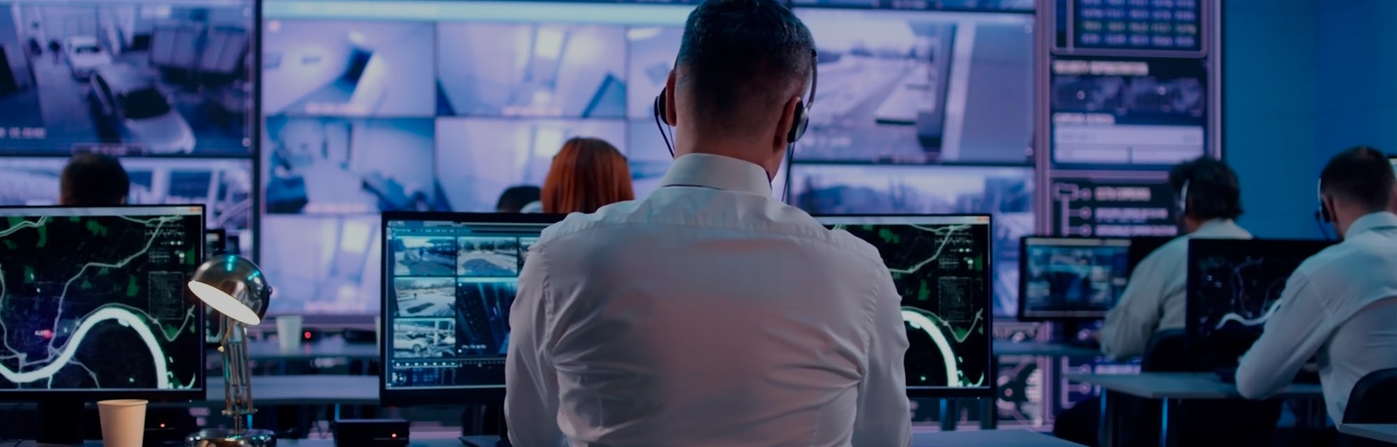 Back view of young man with headset watching surveillance footage and map on computers and speaking ...