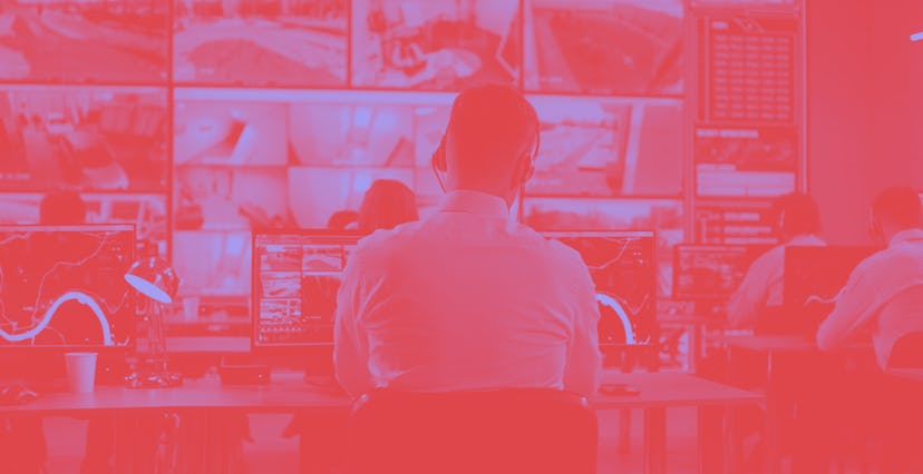 Back view of young man with headset watching surveillance footage and map on computers and speaking ...