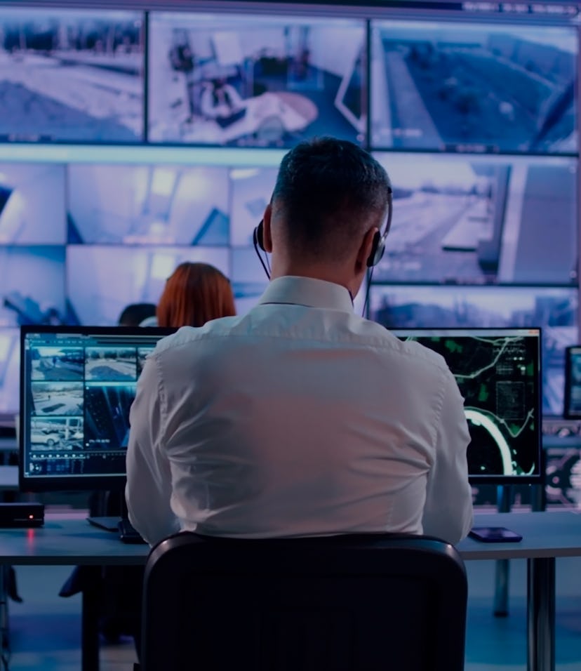Back view of young man with headset watching surveillance footage and map on computers and speaking ...