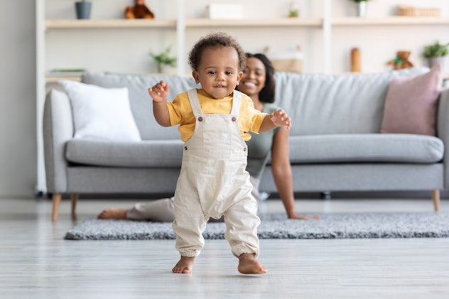Cute black infant boy taking first steps at home, his mother smiling behind him.