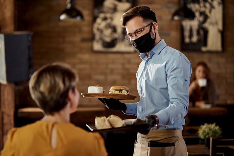 Happy waiter wearing protective face mask and gloves while bringing food to a customer in a pub.