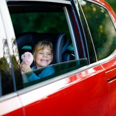 Adorable toddler girl sitting in car seat, holding plush soft toy and looking out of the window on n...