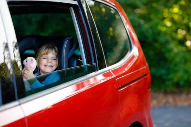 Adorable toddler girl sitting in car seat, holding plush soft toy and looking out of the window on n...