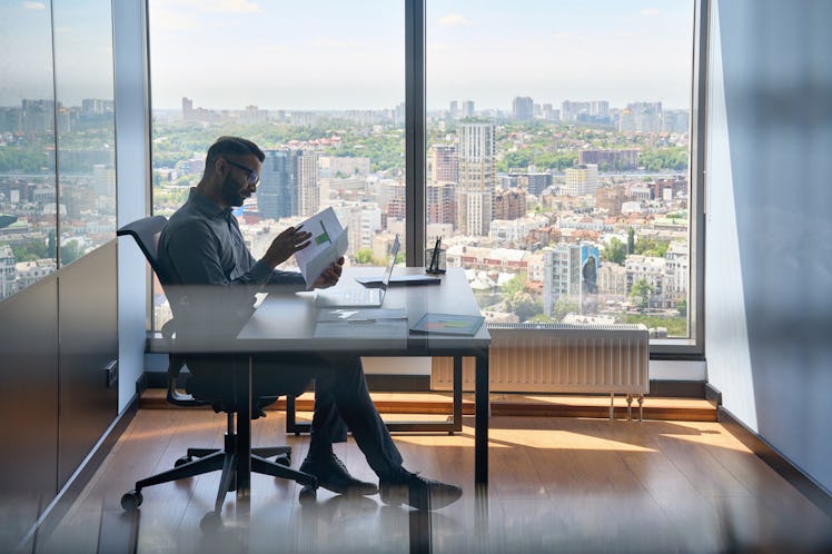 Indian serious investment banker financial analyst sitting at desk with papers working with big data...