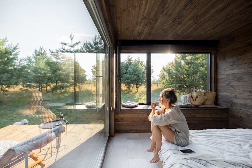 A woman is sitting on the edge of a bed and looking outside towards the woods.