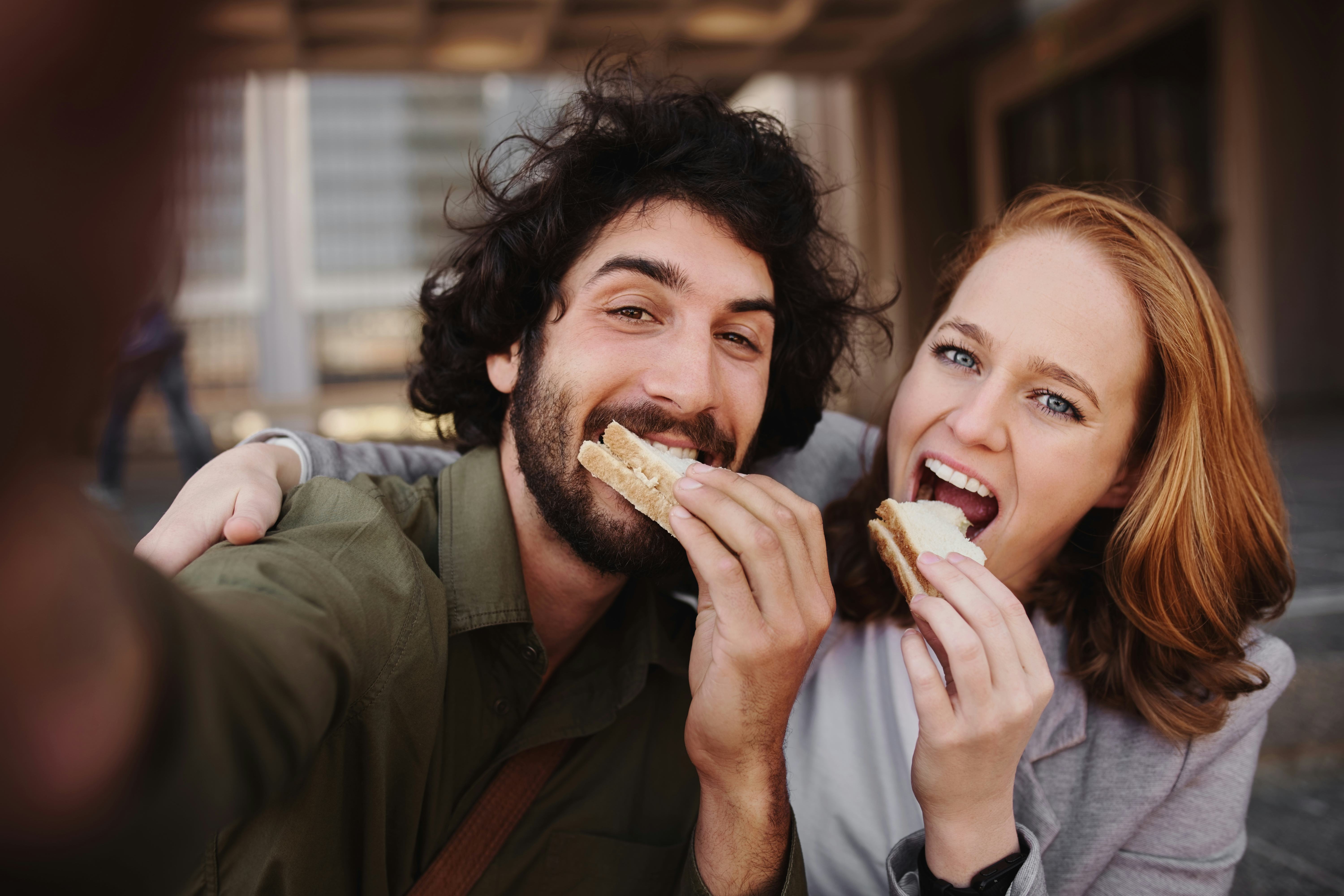 Cheerful young professional couple taking a selfie using smartphone while grabbing a bite of sandwic...