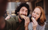 Cheerful young professional couple taking a selfie using smartphone while grabbing a bite of sandwic...