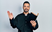 Handsome hispanic priest man with beard holding bible and christian cross smiling with a happy and c...