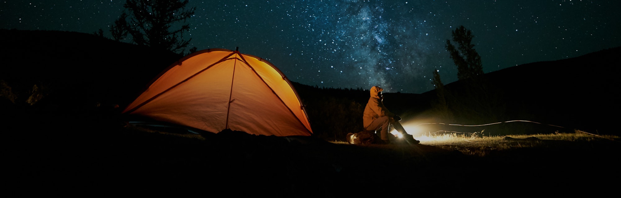 Tourist near his camp tent at night under a sky full of stars. Orange illuminated tent.