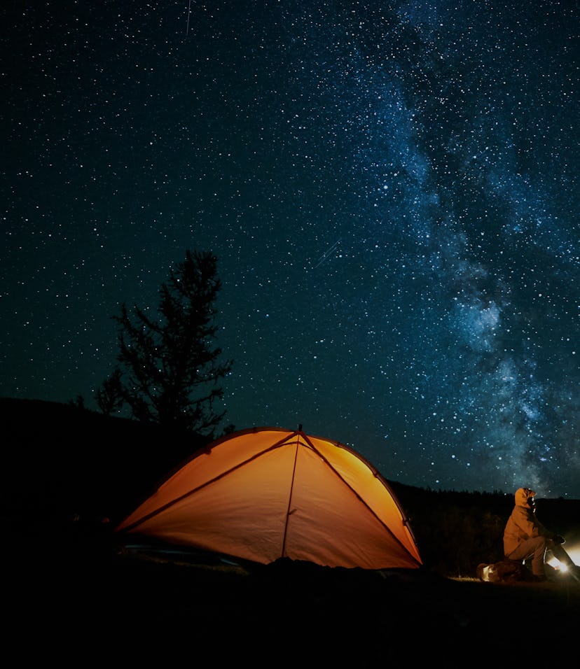 Tourist near his camp tent at night under a sky full of stars. Orange illuminated tent.